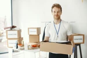 A smiling man carries a donation box while volunteering in a bright office space.